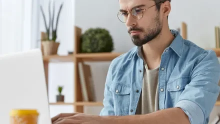 Man with glasses in a denim shirt looking with a serious expression at a laptop screen