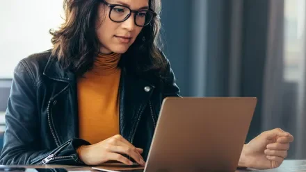 Woman in an orange shirt, black jacket and glasses looking at a laptop screen